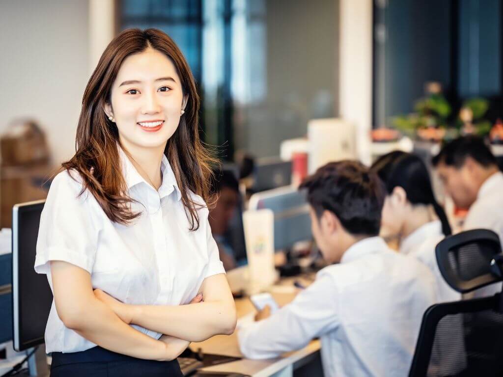 an office lady posing in front of a pc with her colleagues working in the background